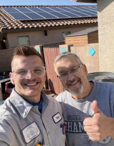 A smiling Olive Air & Heating technician, wearing safety glasses and a uniform with the company logo, takes a selfie with a happy customer giving a thumbs-up. They are standing outdoors near an air conditioning unit, with a home featuring solar panels in the background. The bright sunlight and friendly expressions highlight a successful HVAC installation visit.