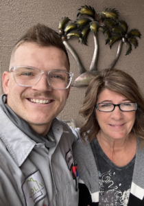 An Olive Air & Heating technician smiles alongside a satisfied customer after completing an HVAC tune-up. The technician is wearing a uniform with the company logo, and the customer is happily posing in front of a decorative palm tree wall art.