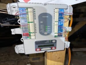 An HVAC zoning control board with multiple connected wires, mounted on a wooden beam in an attic, during an HVAC tune-up.