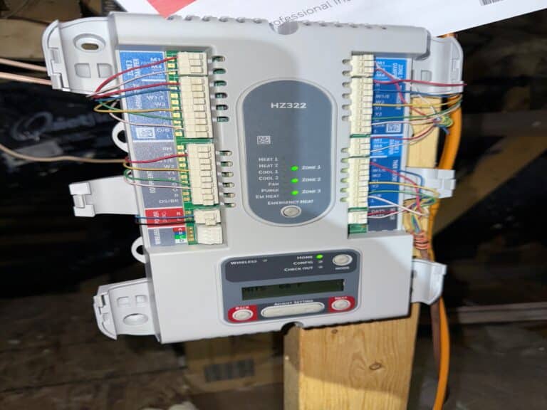 An HVAC zoning control board with multiple connected wires, mounted on a wooden beam in an attic, during an HVAC tune up.